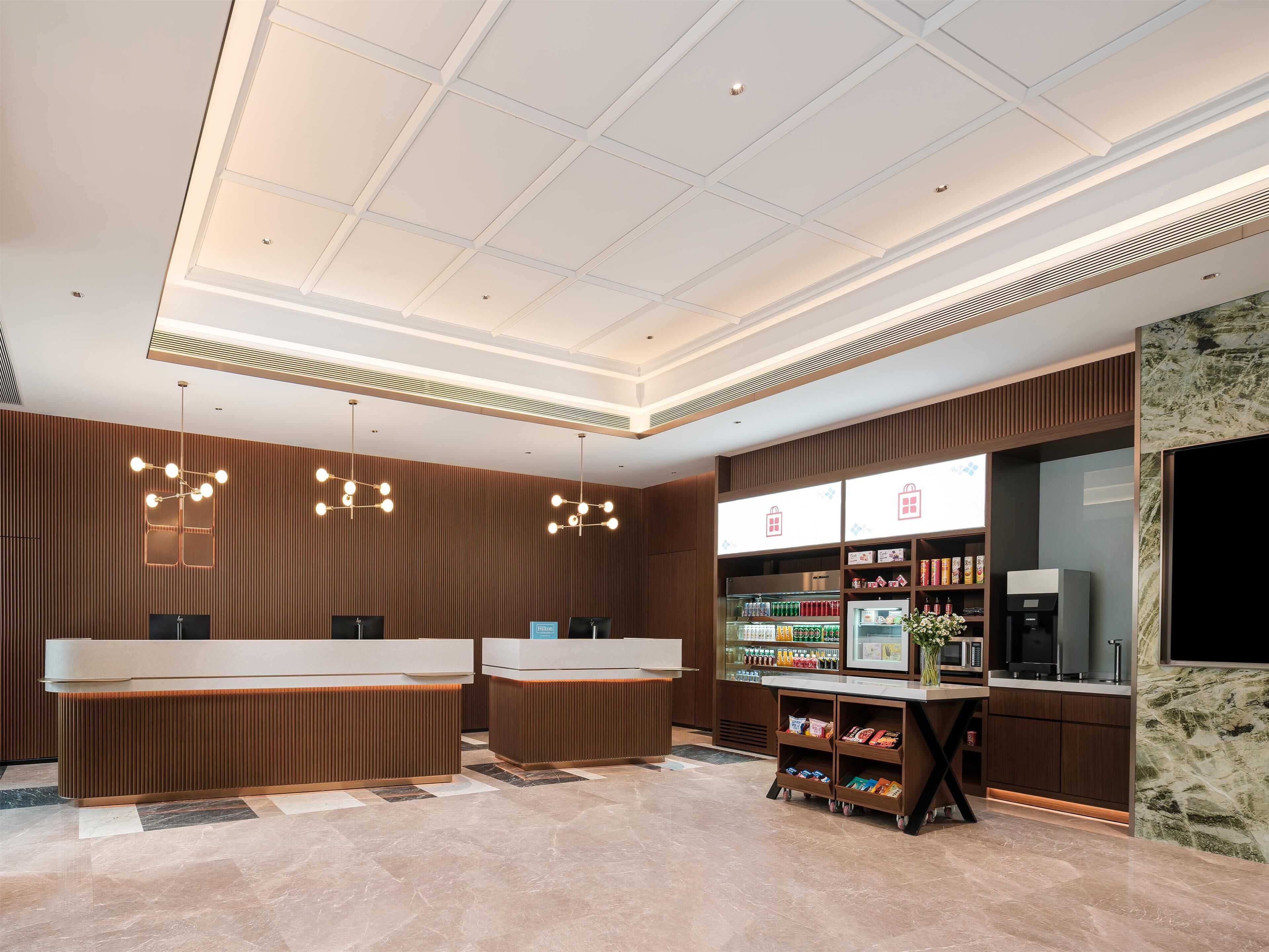A hotel reception area with a modern design, featuring a reception desk, a self-service kiosk, and a refrigerated display case with beverages.