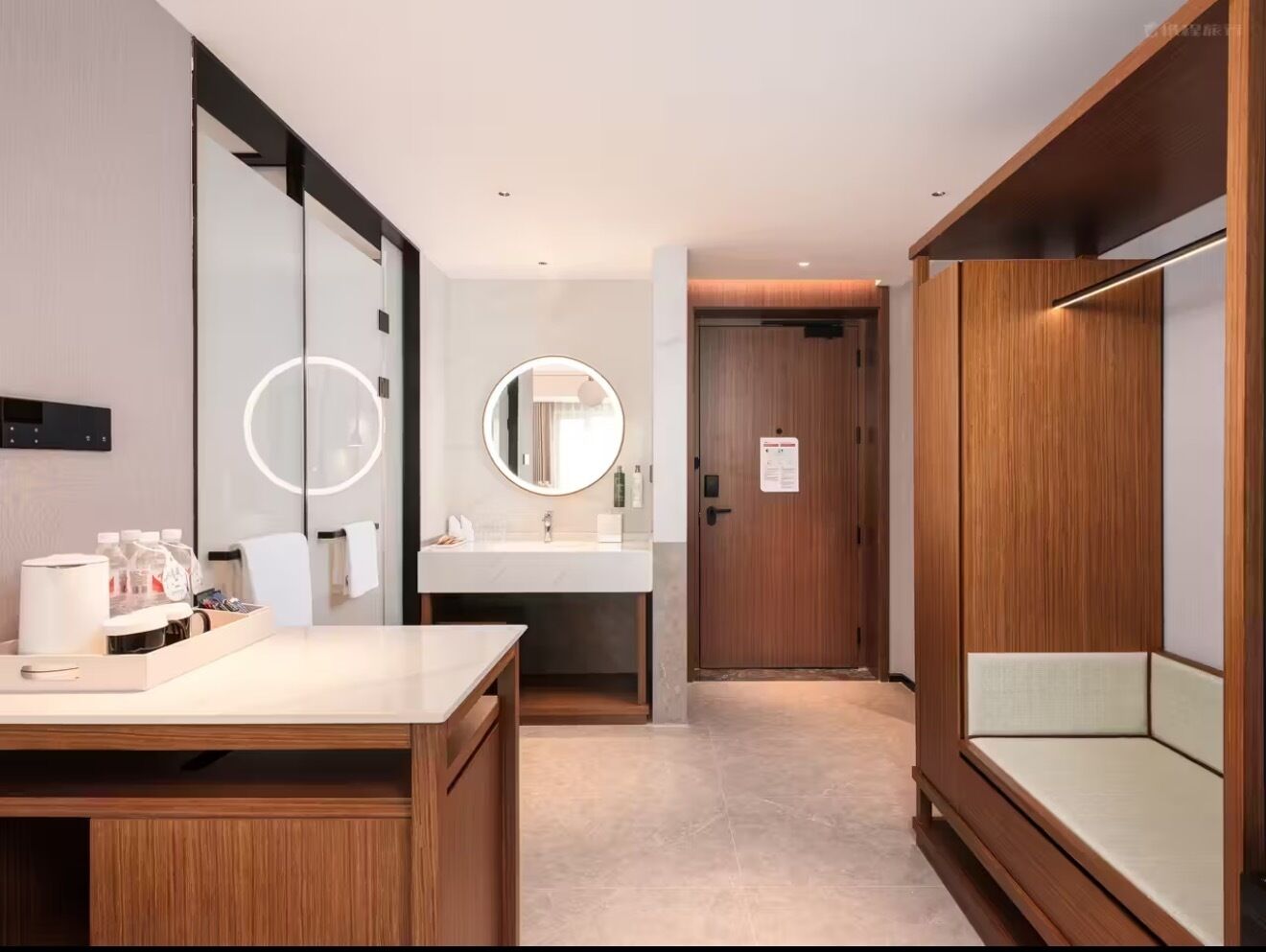 A hotel room with a wooden desk, a round mirror, a white sink, and a wooden wardrobe.