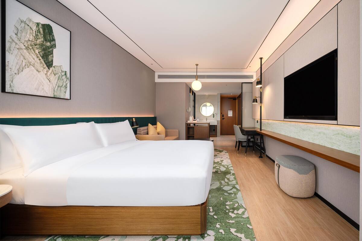A modern hotel room with a large bed, a desk, a chair, a vanity, and a television.