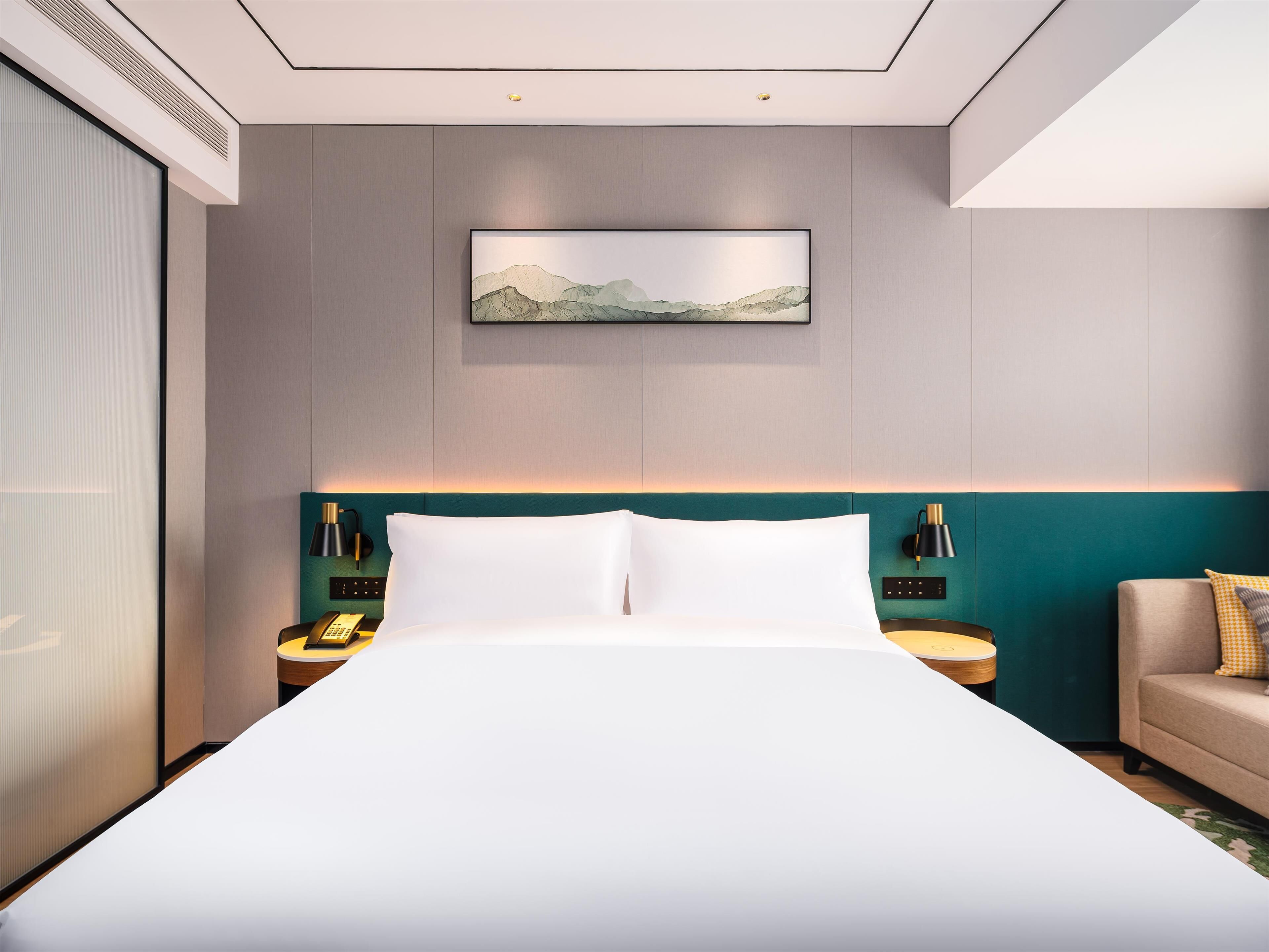 Una habitación de hotel moderna con una cama grande, una pared de acento color verde azulado, un cuadro de paisaje y una mesita de noche con una lámpara.