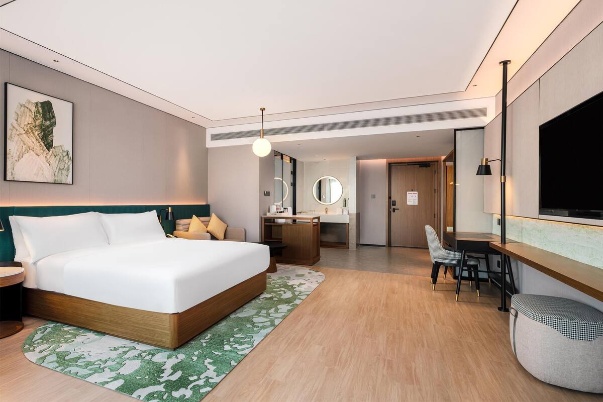A modern hotel room with a large bed, a desk, a chair, a mirror, and a TV.