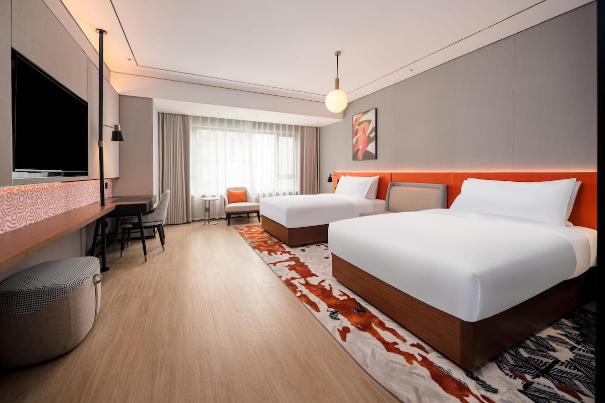 A hotel room with two beds, a desk, a chair, a TV, and a large window with curtains.