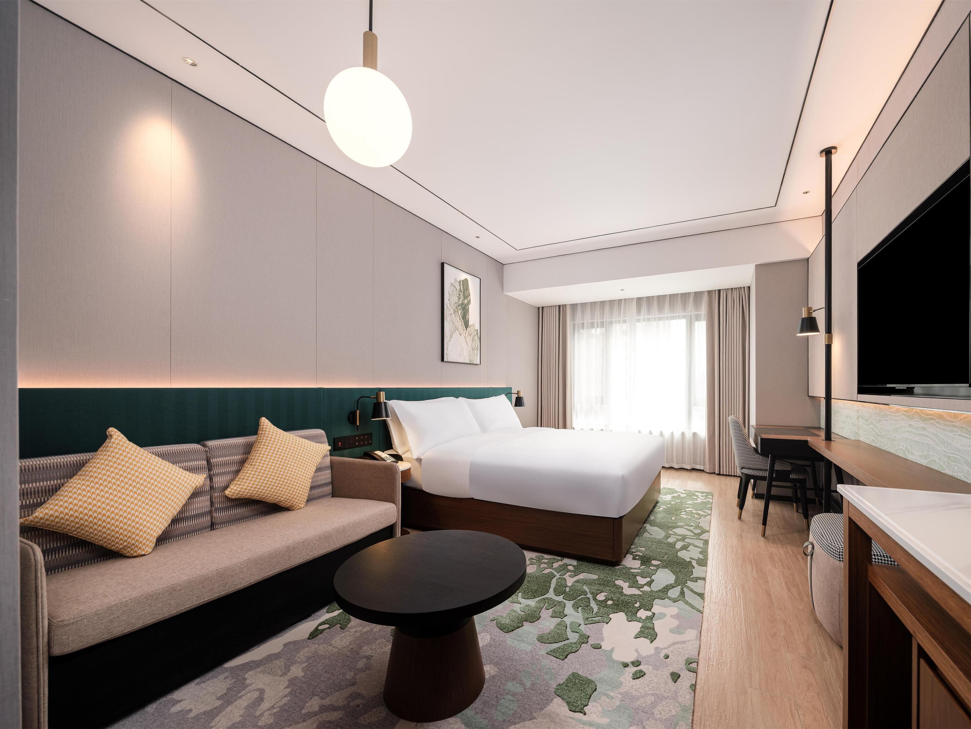 A modern hotel room with a bed, sofa, desk, and TV.