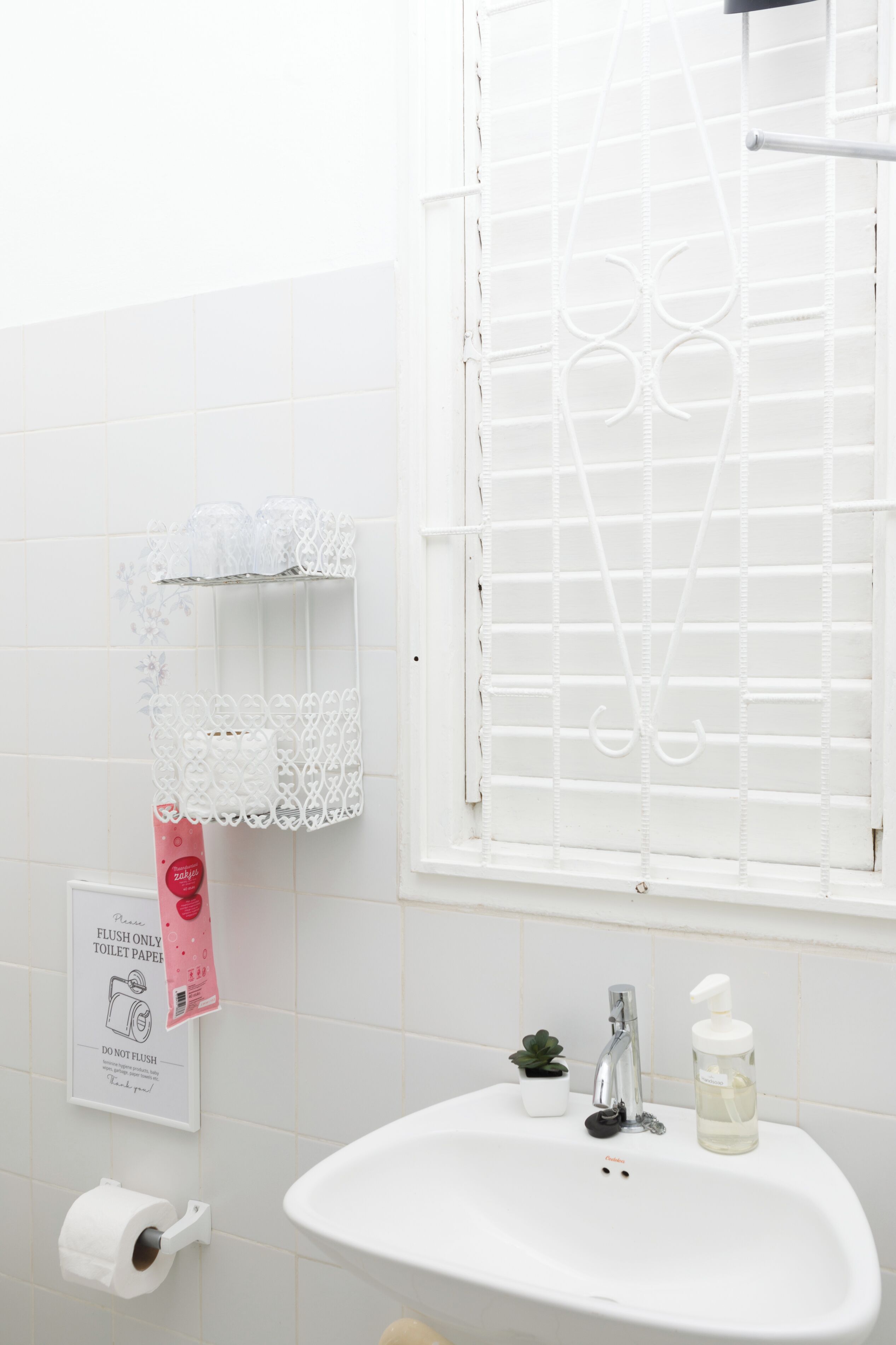 Ein Badezimmer mit einem weißen Waschbecken, einem Toilettenpapierhalter, einer kleinen Topfpflanze und einem Fenster mit Jalousien.