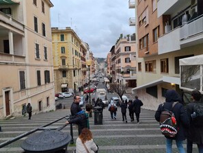 A street with buildings on either side, a staircase leading up, and people walking. - Candia 66 at the Vatican Museums (Roma)