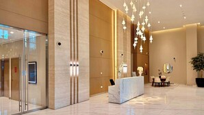Un moderno vestíbulo de hotel con un mostrador de recepción de mármol, una entrada por puerta de cristal y un área de comedor con una planta grande en maceta.