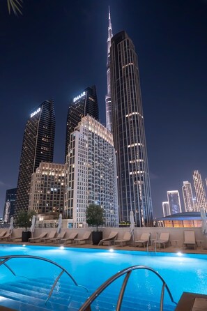A nighttime cityscape with illuminated skyscrapers and a swimming pool area with lounge chairs and umbrellas. - Luxurious 3BR, Burj Khalifa & full fountain view (Dubai)