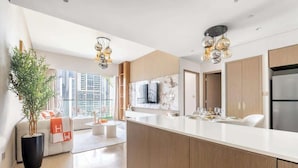 A modern kitchen with a large island, wooden cabinets, and a built-in oven. There is a dining area with a white sofa and a large window offering a view of the city.