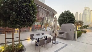 An outdoor seating area with tables and chairs, surrounded by greenery and overlooking a cityscape with tall buildings.