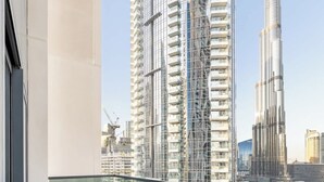 Een balkon met kunstgras, rieten meubilair en een glazen hekwerk met uitzicht op een stadsgezicht met hoge gebouwen.