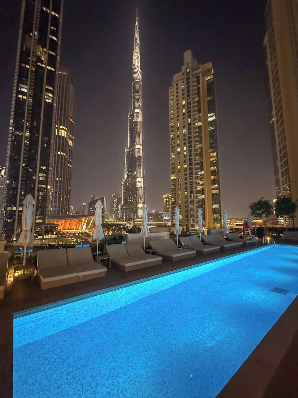 A rooftop pool with lounge chairs and umbrellas, overlooking a cityscape with high-rise buildings, including a prominent tower.