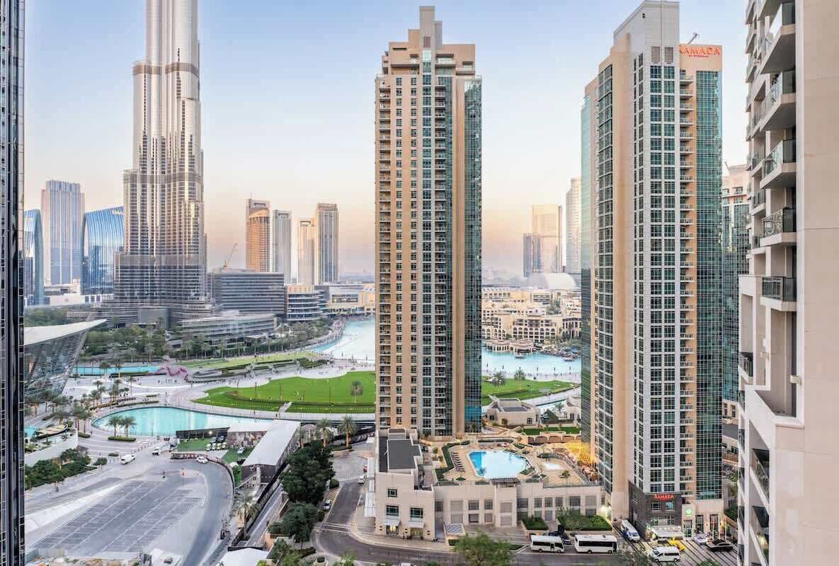 A high-rise building with a modern design, surrounded by other tall buildings, a parking area, and a landscaped area with a pool.