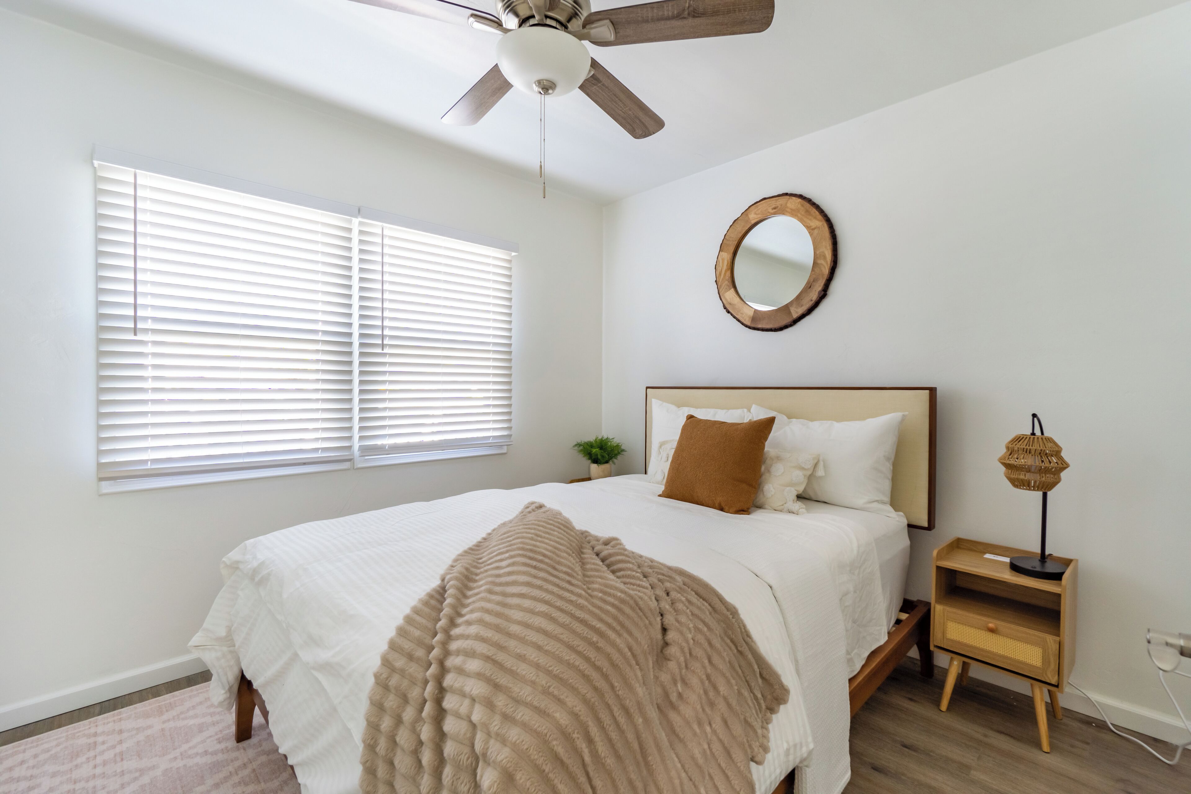 A bedroom with a bed, a ceiling fan, a bedside table with a lamp, and a window with blinds.