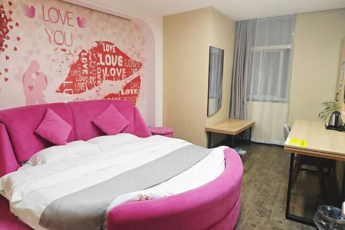 A hotel room with a large bed, a small table, a chair, a mirror, and a wall with a love-themed poster.