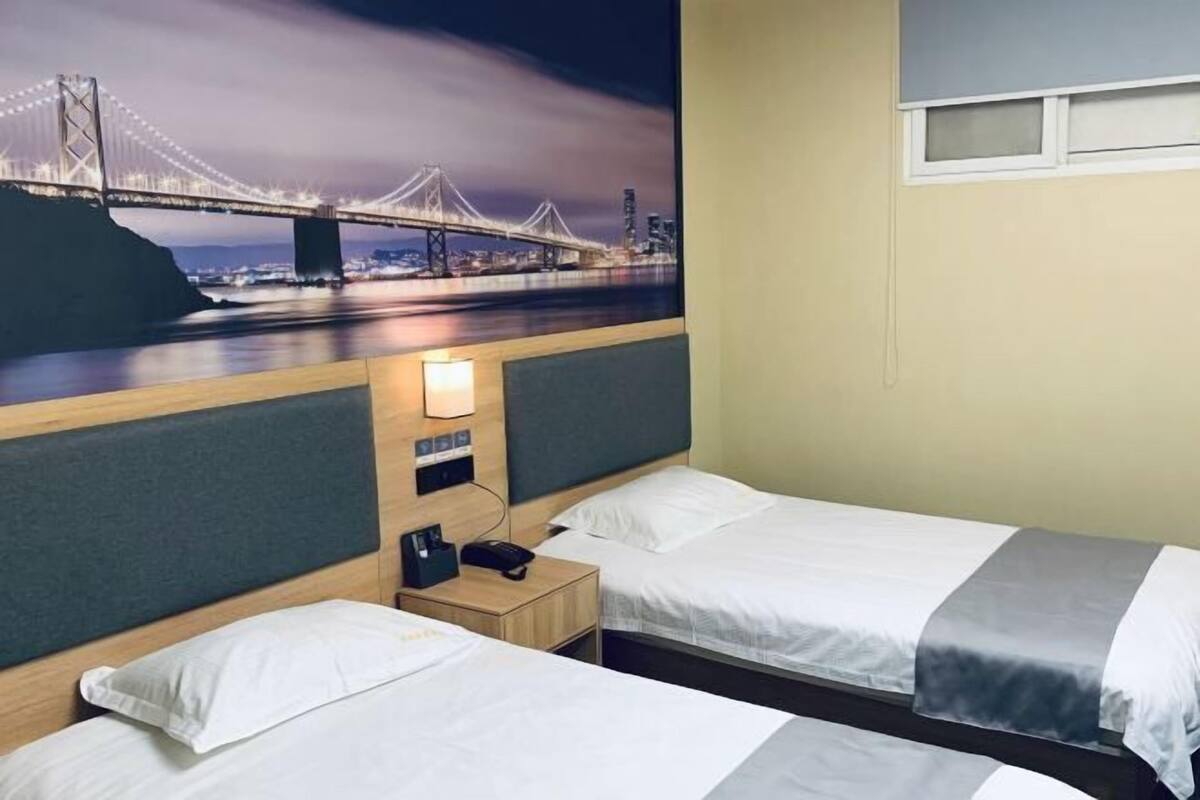 A hotel room with two beds, a nightstand with a lamp, and a wall-mounted TV displaying a nighttime cityscape.