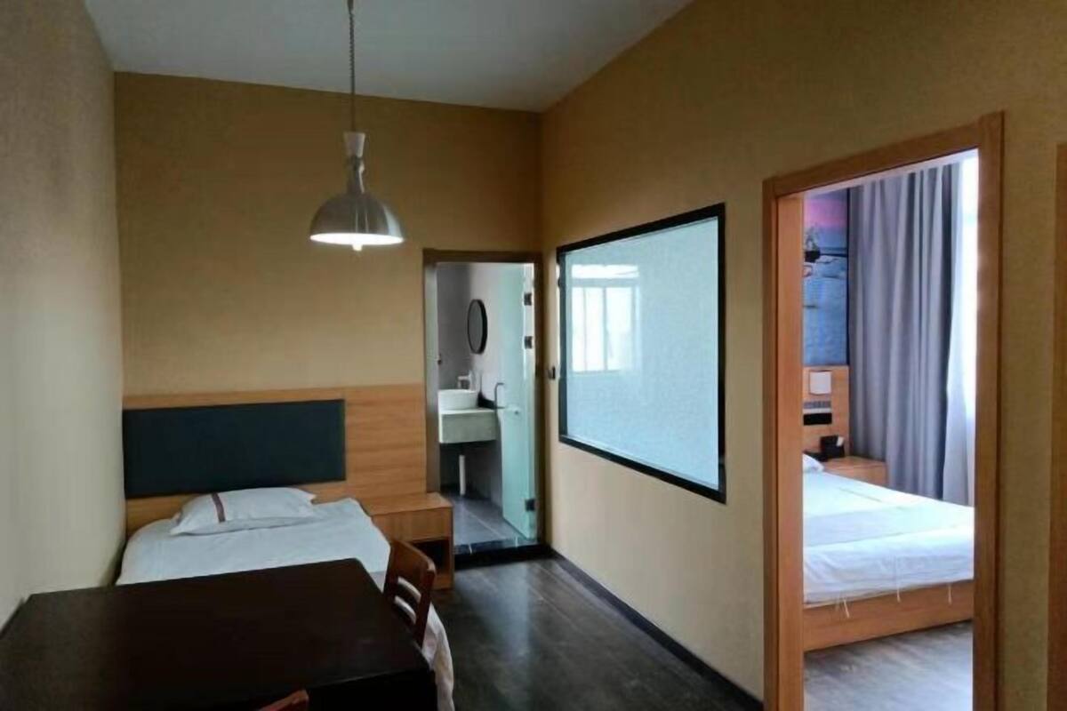 A hotel room with a bed, a desk, a chair, a hanging light, a bathroom visible through an open door, and a large window.