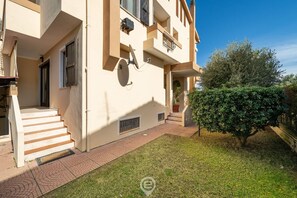 A multi-story building with a staircase leading to the entrance, a small garden area with a tree, and a satellite dish on the wall.