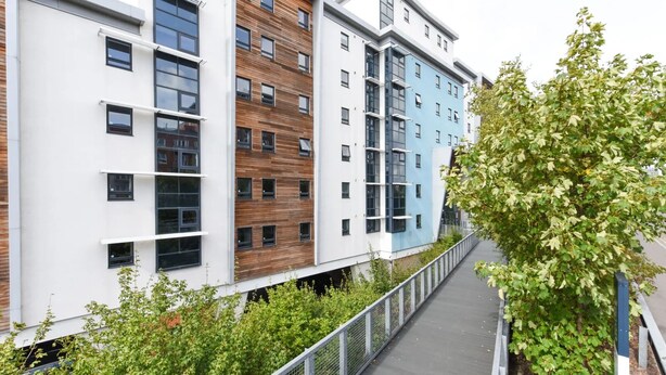 Un edificio moderno de varios pisos con una combinación de fachadas blancas, de madera y azules, un paseo cubierto y abundante vegetación.