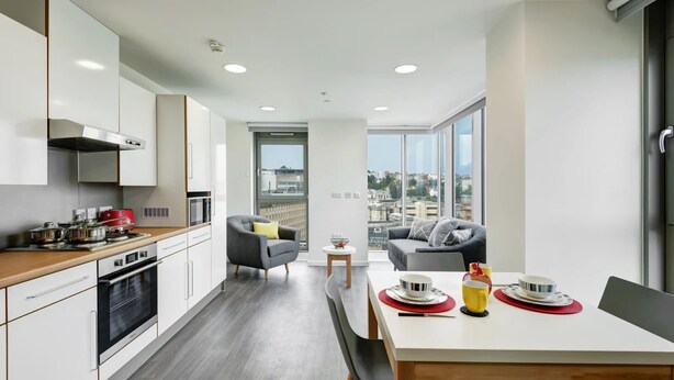 A modern kitchen with a stove, oven, and red appliances. There is a dining area with a table set for two, and a sofa in the background. - New Bridewell (Bristol)