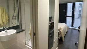 A modern hotel room with a glass shower enclosure, a sink, a bed with a white duvet, and a window providing a view of the outside. - New Bridewell (Bristol)