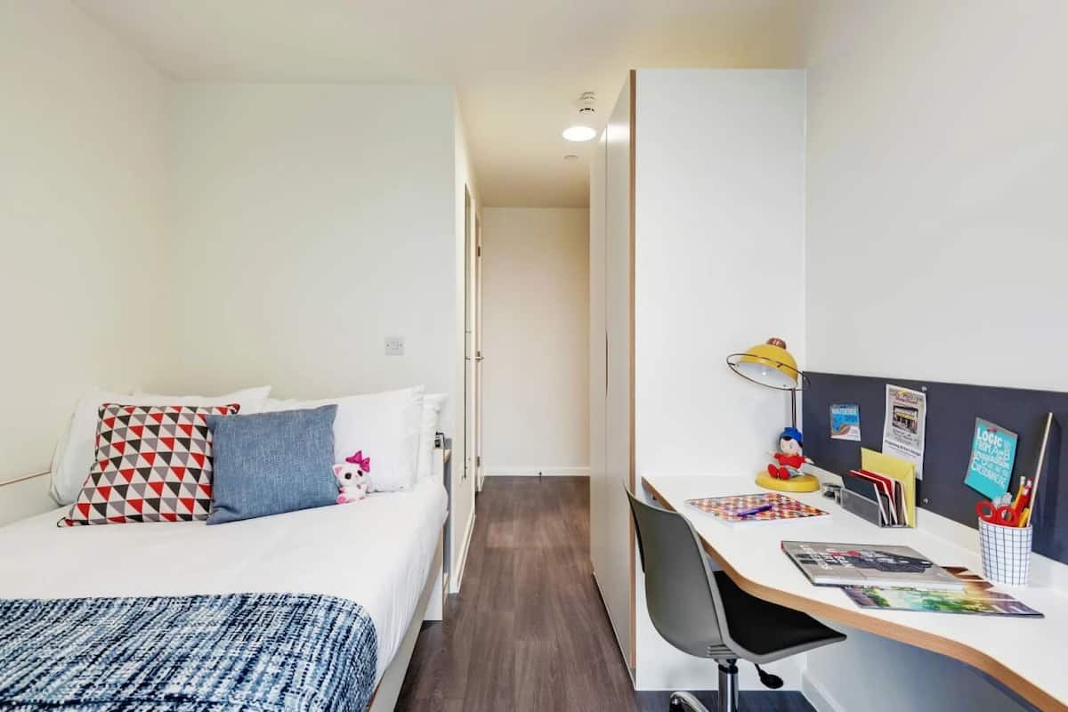 A hotel room with a bed, a desk, and a chair. There is a lamp on the desk, a small toy on the bed, and a bulletin board with notes and pictures.