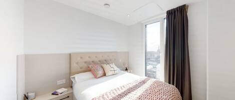 Une chambre d’hôtel moderne, dotée d’un grand lit, d’une table de chevet, d’une commode et offrant une vue sur la ville grâce à une grande fenêtre.