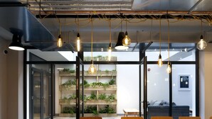 A modern meeting room with a wooden table, chairs, hanging pendant lights, and a glass wall with a vertical garden. - For Students Only Abodus The Walls Southampton - Campus Accommodation (Southampton)