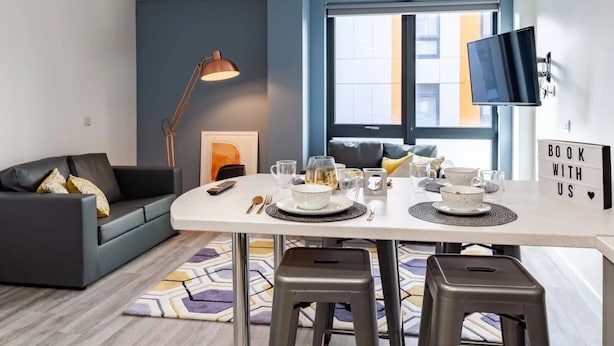 A modern living room with a dark grey sofa, a white dining table set with plates, glasses, and cutlery, a floor lamp, and a TV mounted on the wall. - Student Only Oldway Centre (Swansea)