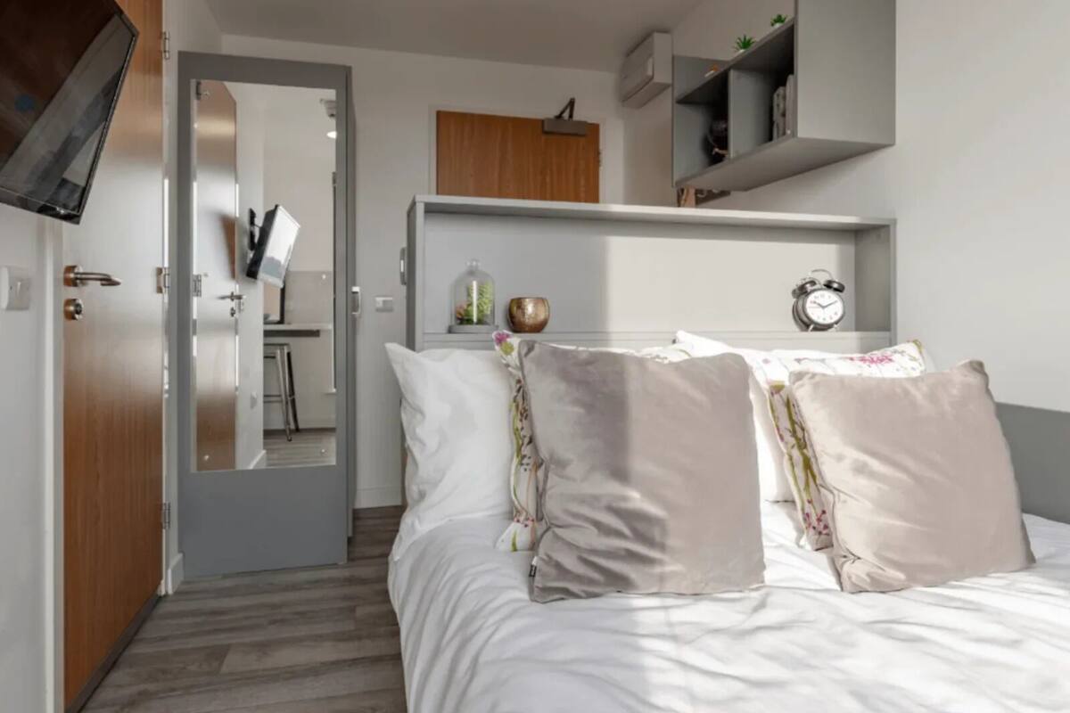 A bedroom with a bed, a mirror, a desk, and a shelf with a clock.