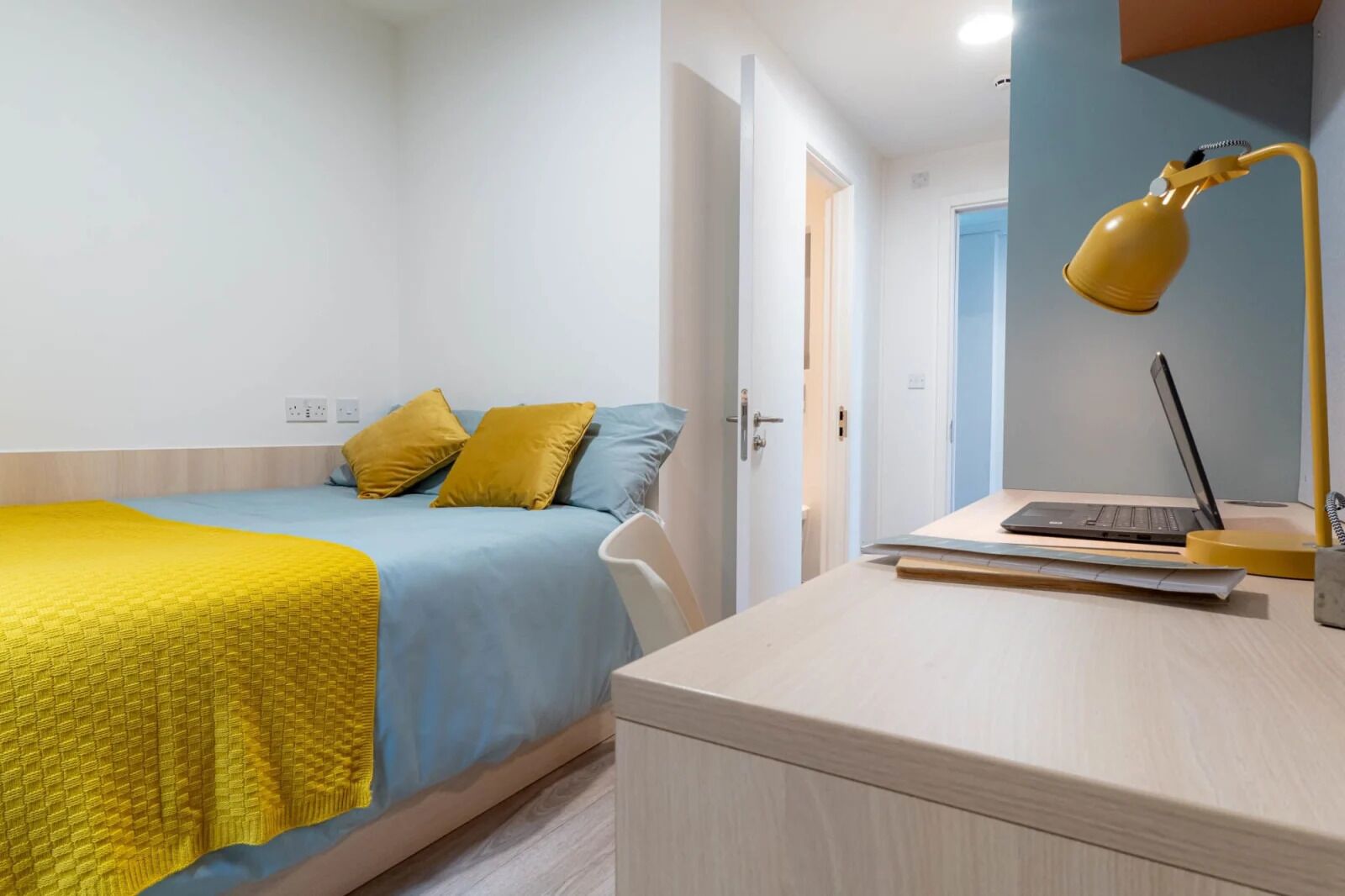 A bedroom with a bed, a desk with a laptop, and a yellow desk lamp.
