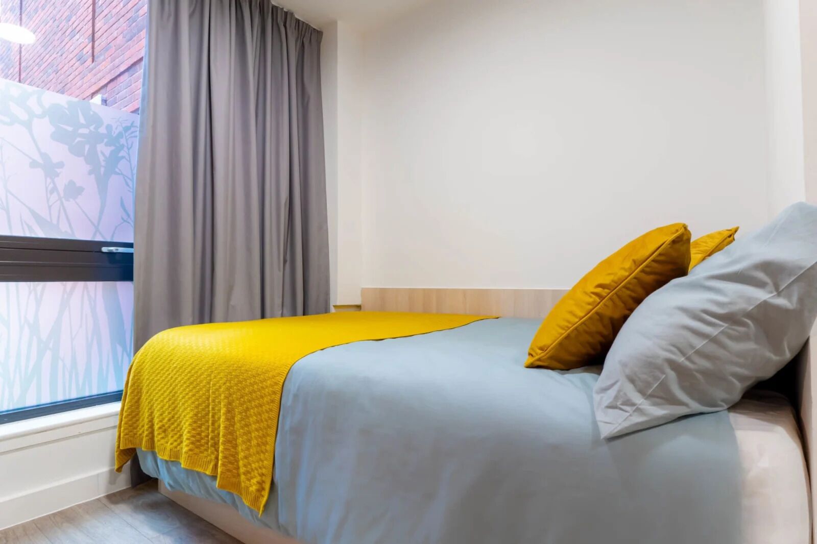 A bedroom with a bed, a window with curtains, and a view of a brick building.