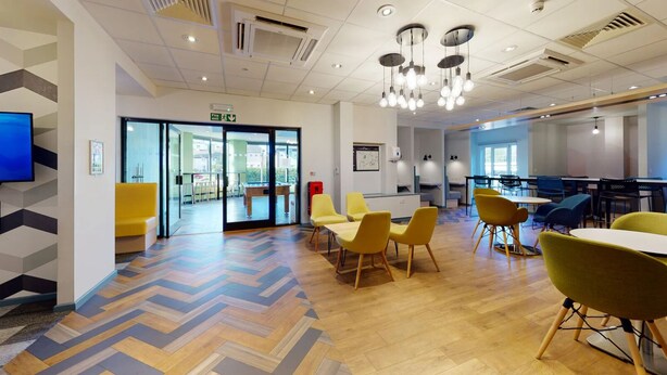 A modern interior with a herringbone wood floor, yellow and green chairs, a fire exit sign, and a flat-screen TV on the wall. - Opal Court (Leicester)