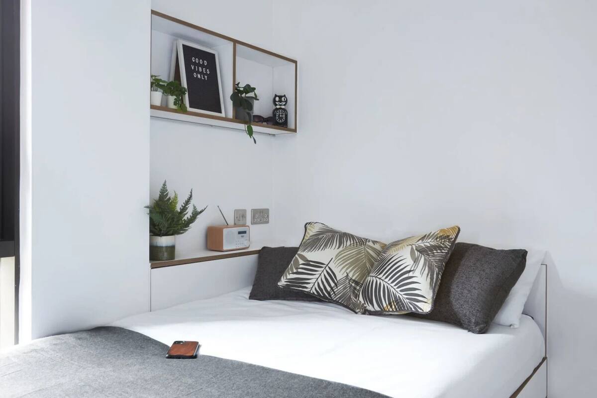 A modern bedroom with a white bed, a grey throw blanket, a wooden bedside table with a radio, a wall-mounted shelf with plants and a framed sign, and a grey pillow.