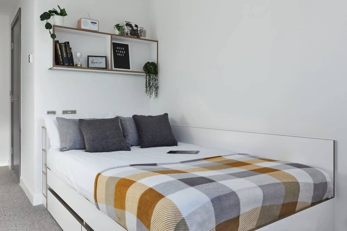 A modern bedroom with a bed, a bedside table, a shelf with books and a plant, and a wall-mounted radio.