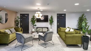 Un salon moderne avec deux canapés verts, une table basse ronde et des fauteuils gris. Un grand téléviseur est fixé au mur, et deux portes sont étiquetées « Salle des plantes » et « Salle des communications ».