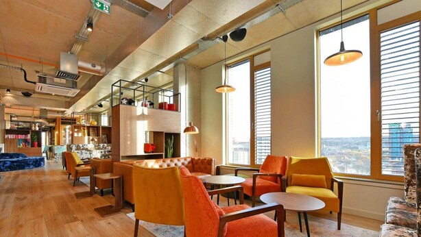 A modern lounge area with wooden flooring, orange and yellow seating, and large windows offering a city view. - For Student Only Liv Student Sheffield - Campus Accommodation (Sheffield)