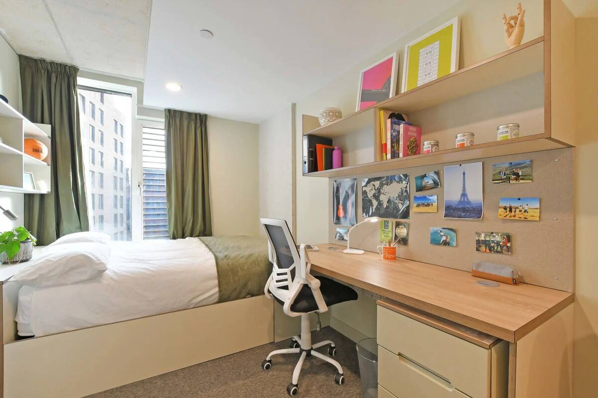 A hotel room with a bed, desk, chair, and shelves with books and photos.
