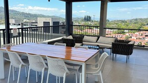 An outdoor patio with a wooden table and white chairs, a sofa, and a view of a cityscape and hills. - Modern Apartment with Volcano View 901 Santa Tecla (Santa Tecla)