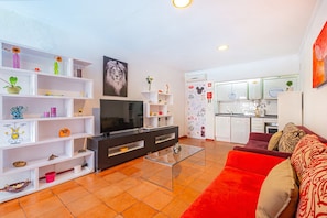 A modern living room with a flat-screen TV, a glass coffee table, and a red sofa. There is a kitchen area with white cabinets and a tiled backsplash.