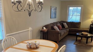 A dining area with a round table and chairs, a chandelier, a sofa, and a window with blinds. - Charming 3-Bedroom Retreat with Waterway Access in Hampstead, NC. Hideaway Haven (Hampstead)