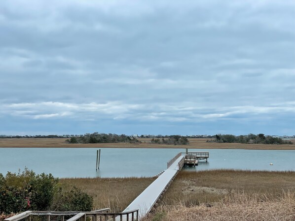 Une longue jetée en bois s’étendant dans une étendue d’eau calme, entourée de terres marécageuses et d’une végétation clairsemée.