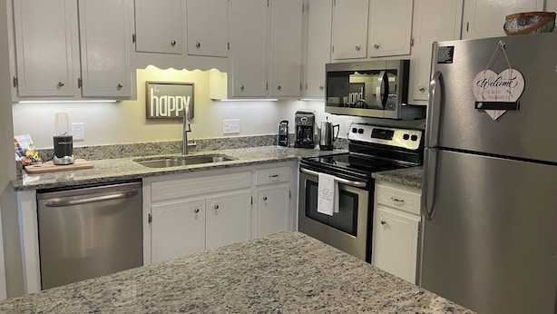 A modern kitchen with white cabinets, granite countertops, stainless steel appliances, and a 'be happy' sign. - Charming 3-Bedroom Retreat with Waterway Access in Hampstead, NC. Hideaway Haven (Hampstead)