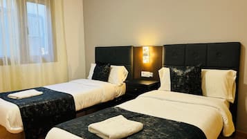 Een hotelkamer met twee bedden, elk met een donker hoofdeinde en witte beddengoed. Tussen de bedden staat een nachtkastje met een lamp en een stopcontact.