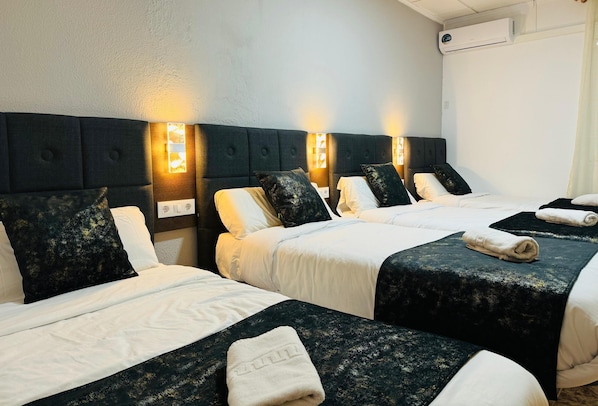 A hotel room with three beds, each with a dark headboard and white bedding. There are wall-mounted lights above each bed, and a white air conditioning unit on the wall. - Ballestar Hotel Bar & Grill (Barajas de Melo)