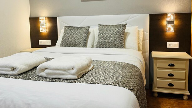 Een net opgemaakt bed met een bedsprei met patroon, twee kussens en opgevouwen handdoeken. Er staat een nachtkastje met een lamp en een wandlamp.