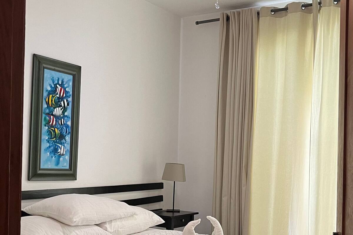 A hotel room with a bed, a nightstand, a lamp, a ceiling fan, a painting, and curtains.