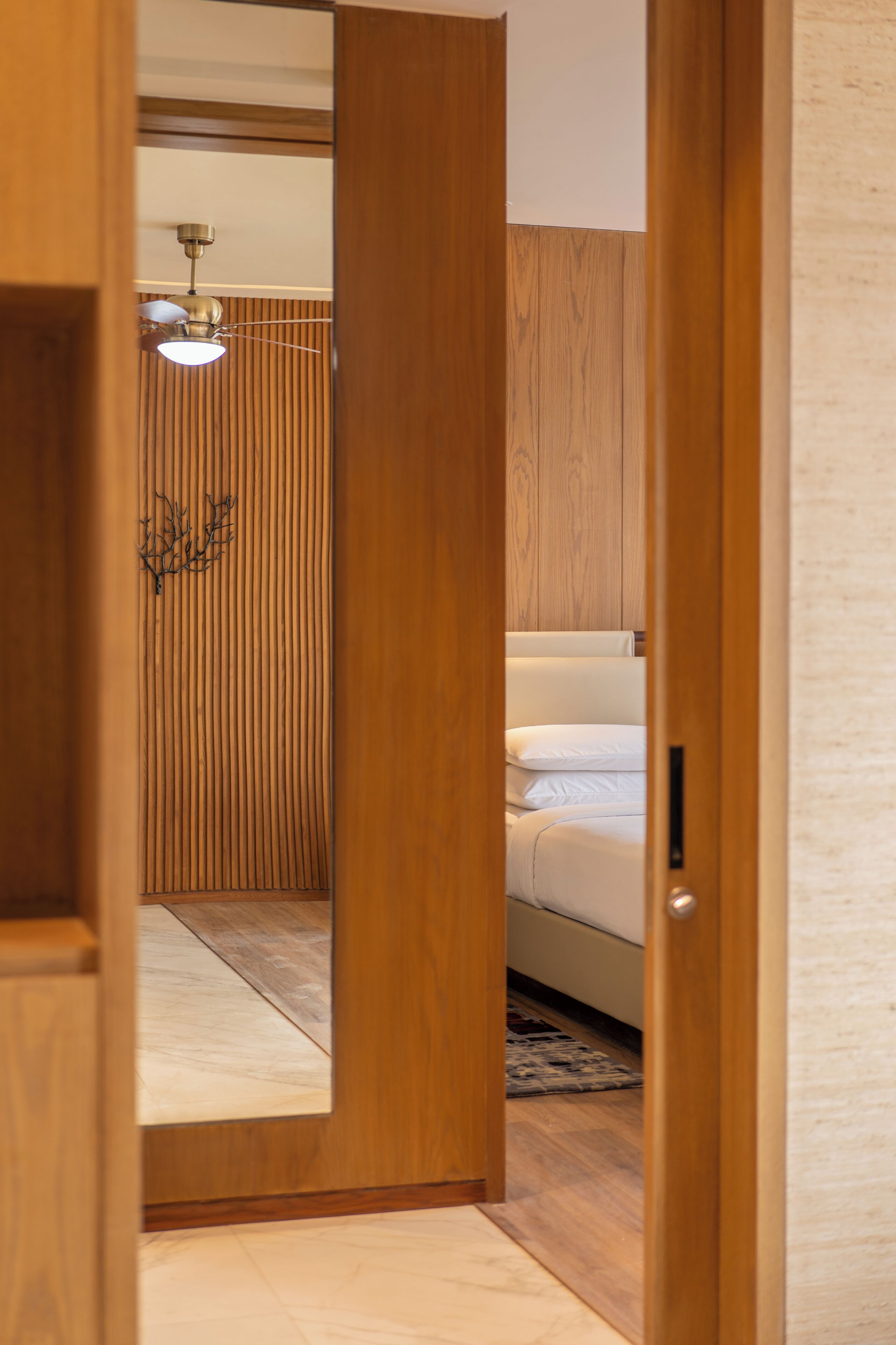 Une porte en bois donne sur une chambre à coucher équipée d’un lit, d’un ventilateur de plafond et d’un luminaire fixé au mur.