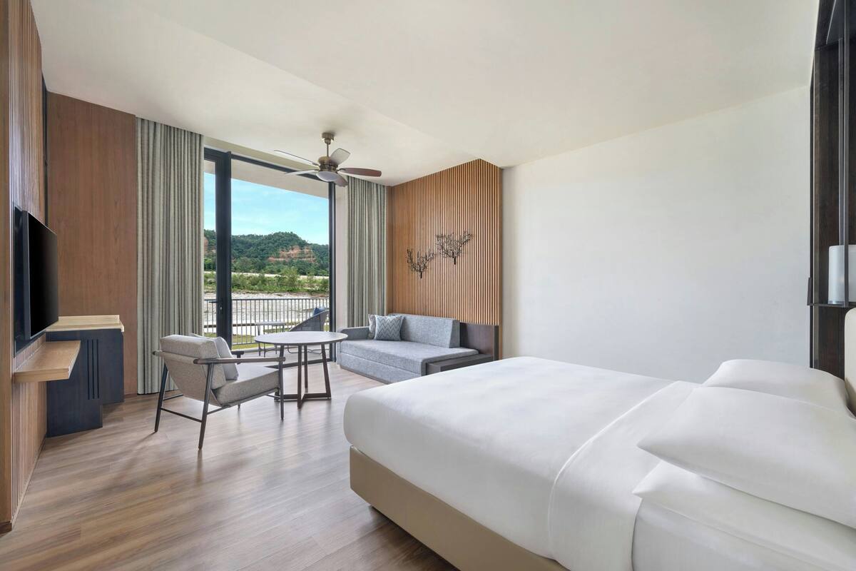 A modern hotel room with a large bed, a desk, a chair, and a balcony with a view of the hills.