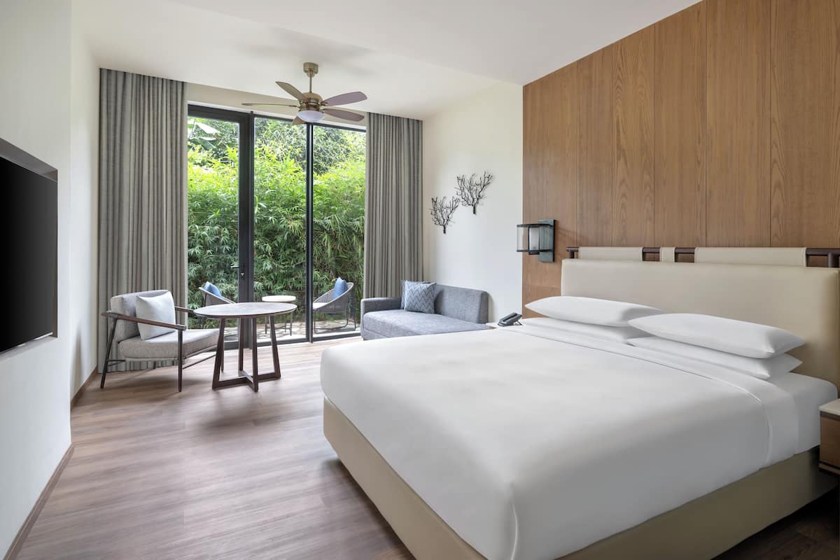 A hotel room with a large bed, a desk with a chair, a sofa, and a view of greenery through the windows.