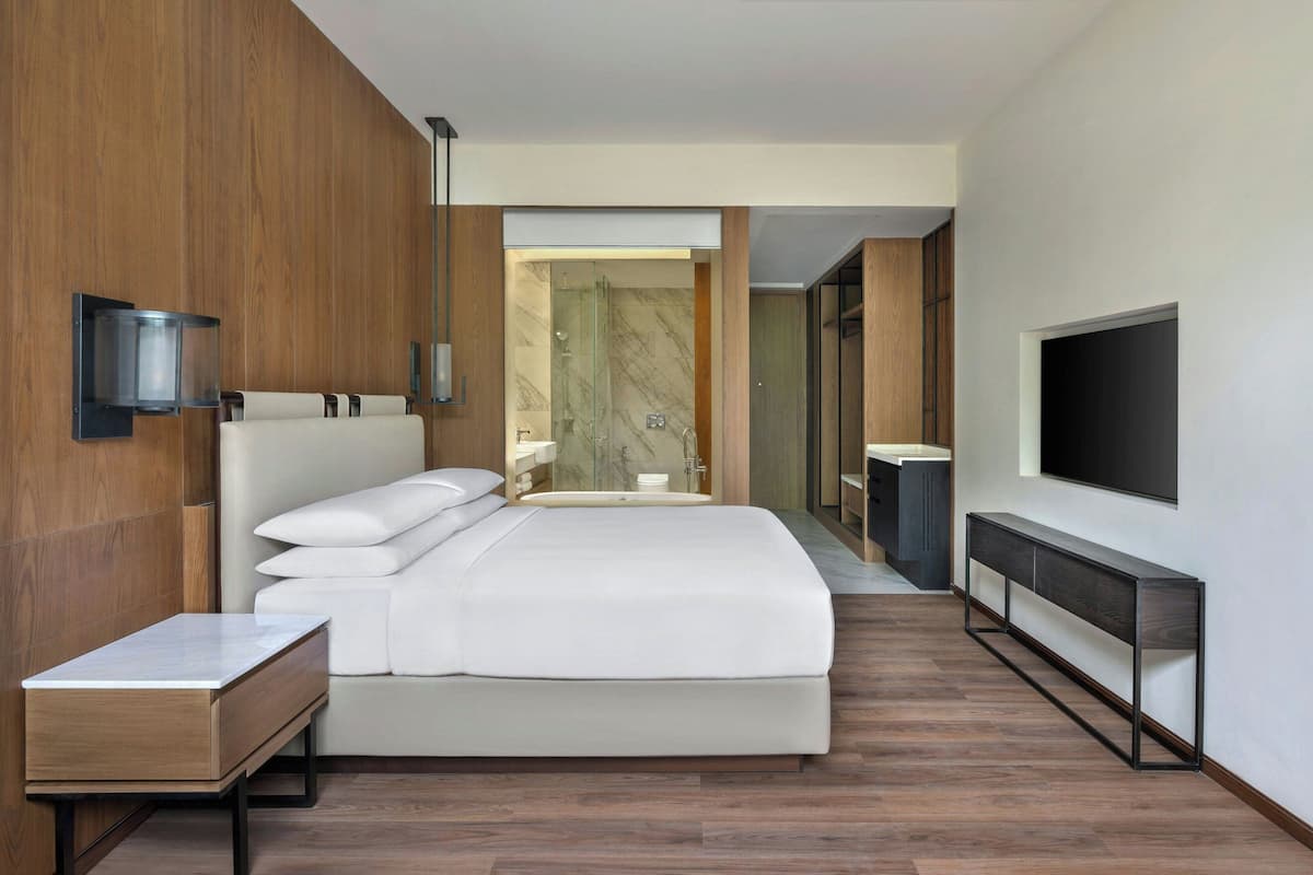 A modern hotel room with a large bed, a bedside table, a TV mounted on the wall, and a bathroom visible through an open door.
